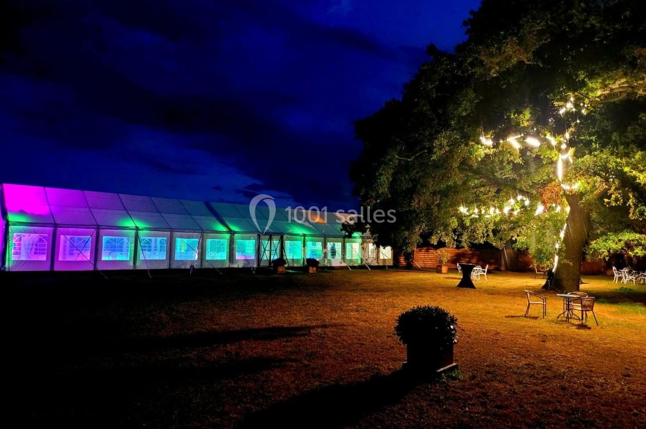Grande tente éclairée de couleurs vives et arbre illuminé dans un espace extérieur en soirée.
