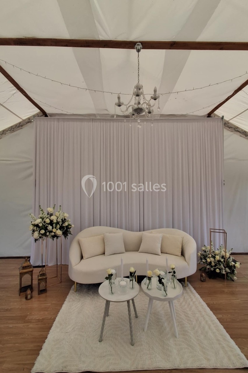 Canapé beige avec coussins, tables basses décorées de fleurs, tapis blanc, rideau et lustre dans une tente élégante.