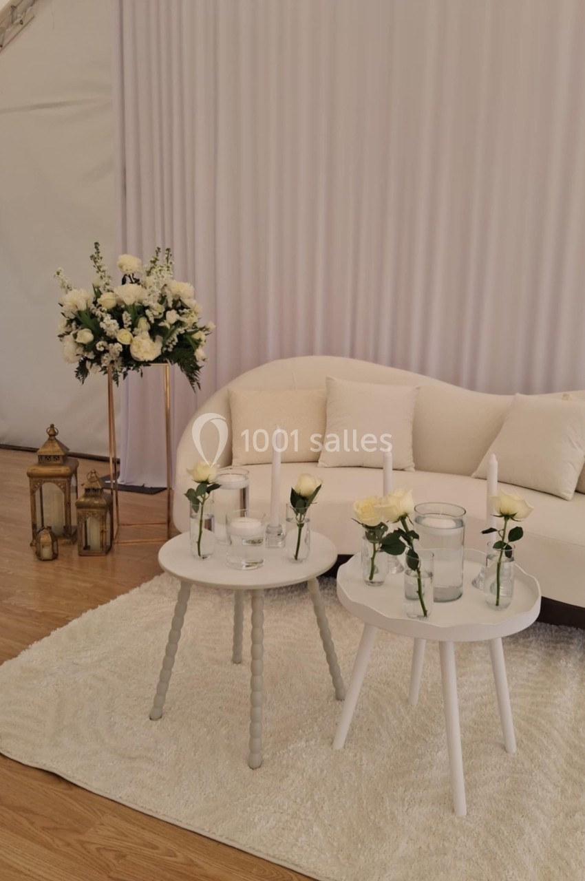 Deux tables blanches décorées de bougies et de roses blanches devant un canapé beige, dans un espace élégant et lumineux.