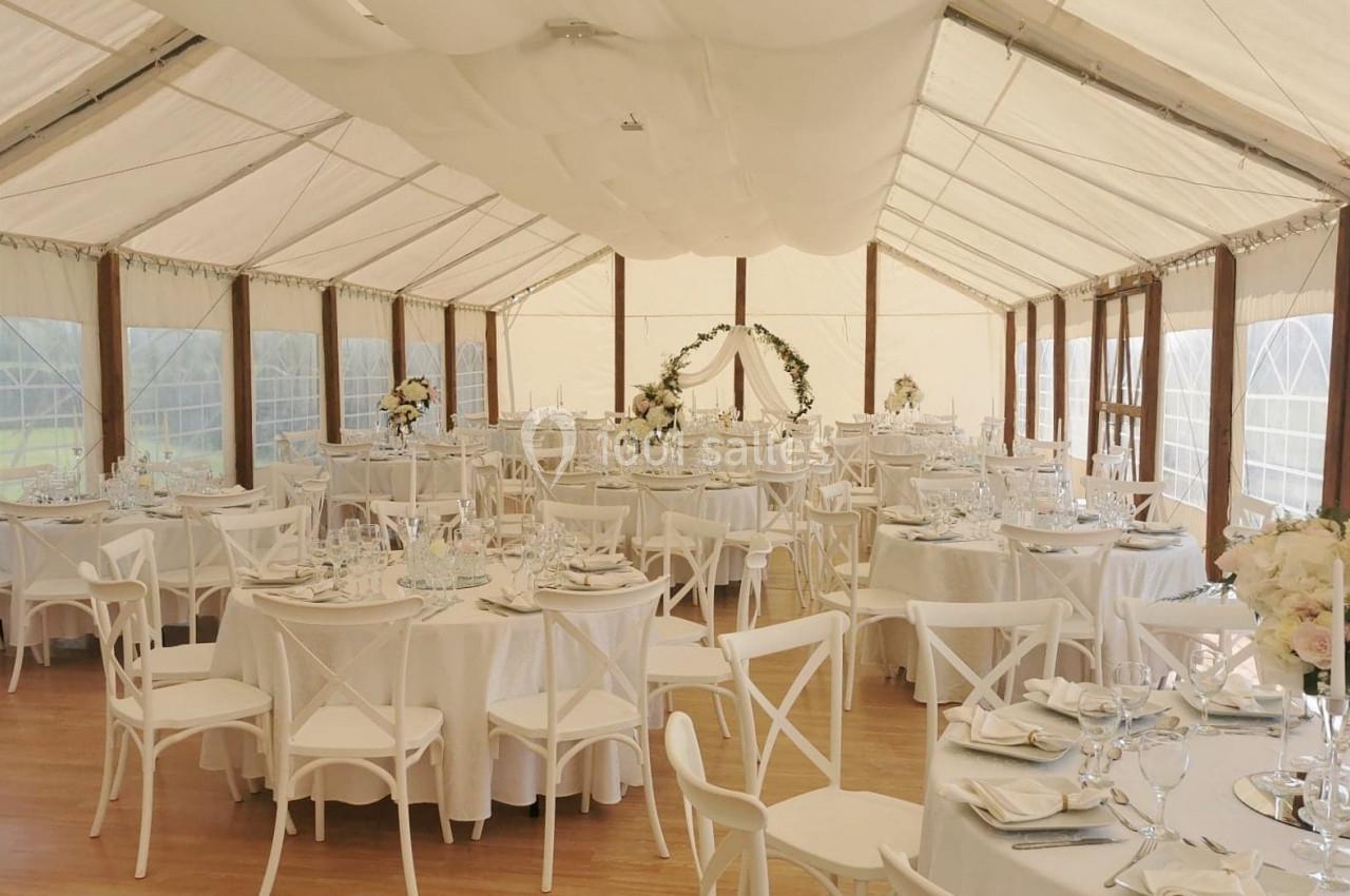 Salle de réception décorée avec des tables rondes, nappes blanches, chaises en bois et arche fleurie au fond.