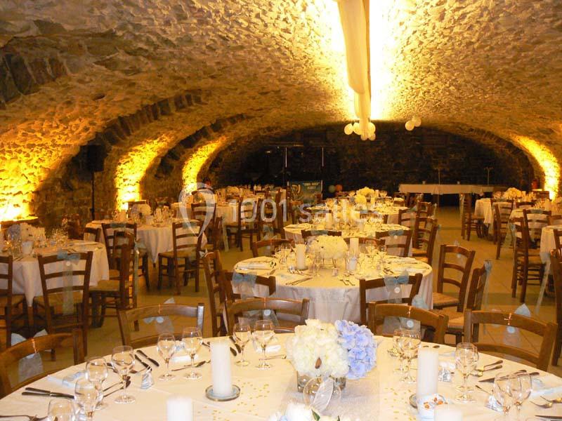 Salle voûtée en pierre éclairée, aménagée avec des tables rondes décorées pour un événement festif.