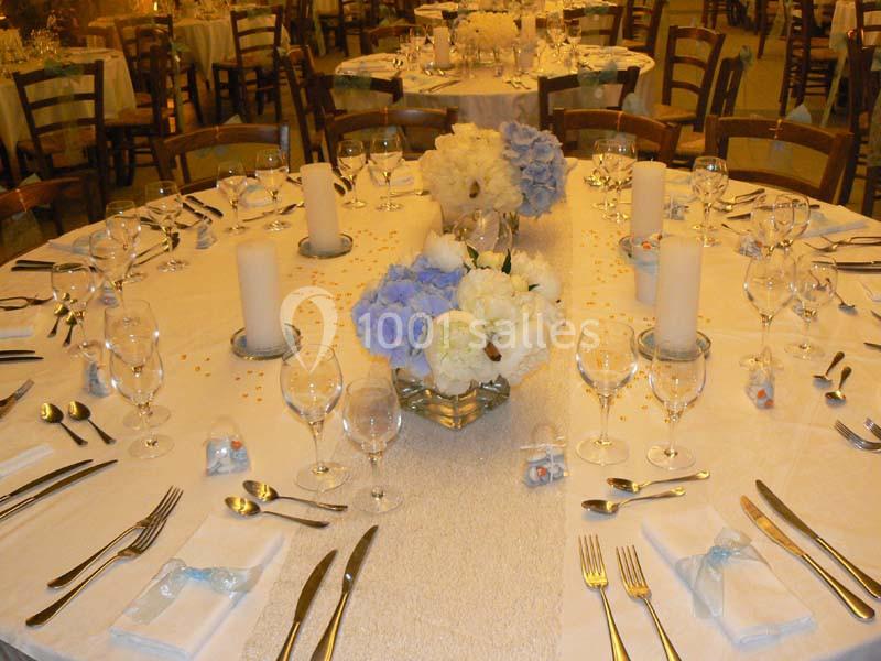 Table ronde élégamment dressée avec nappes blanches, couverts, verres, bougies et bouquets de fleurs bleues et blanches.