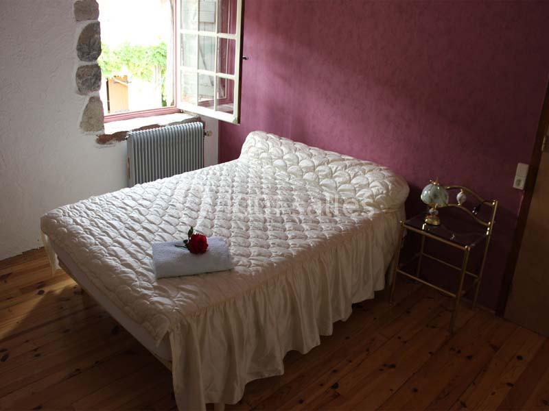 Lit double avec couvre-lit blanc dans une chambre aux murs violets, parquet en bois et fenêtre ouverte sur l'extérieur.