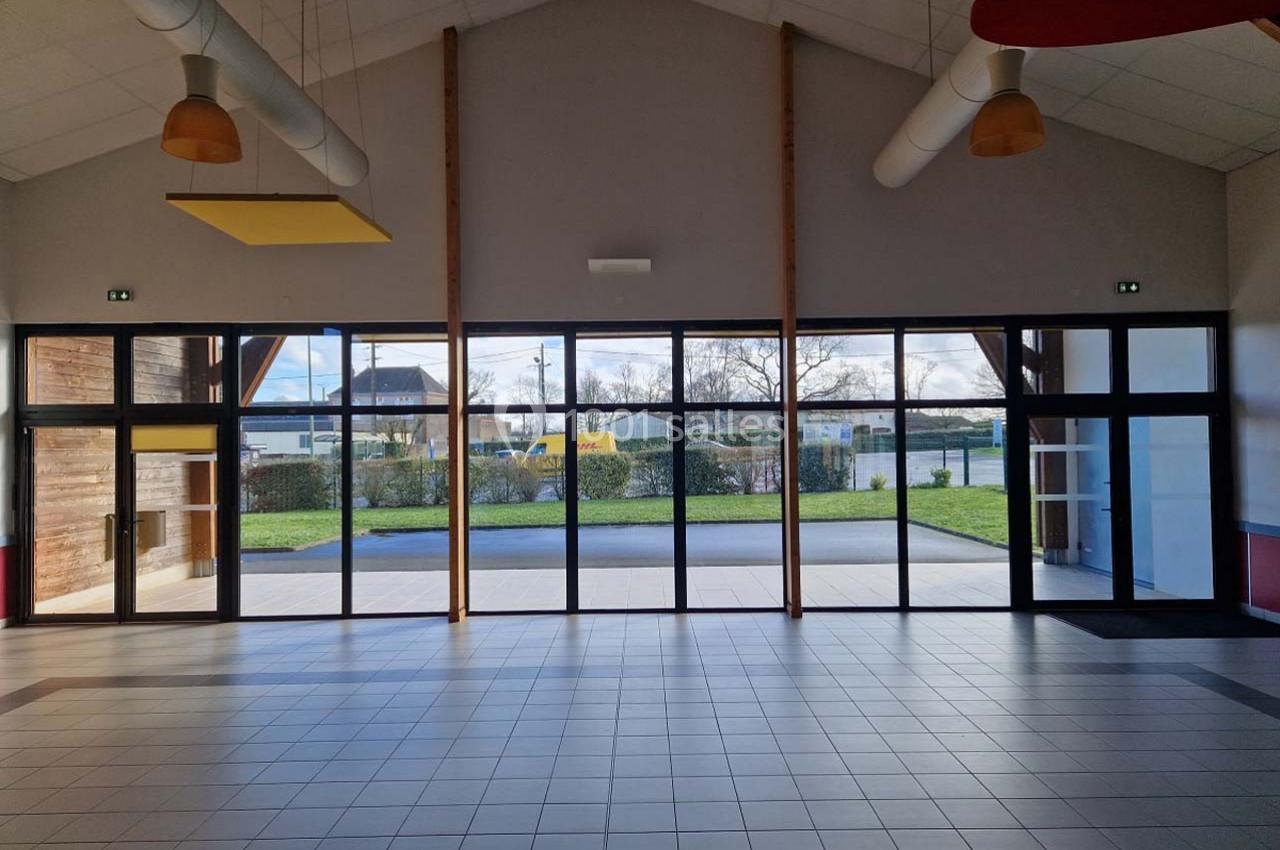 Grande salle lumineuse avec des baies vitrées donnant sur un espace extérieur avec pelouse et route en arrière-plan.