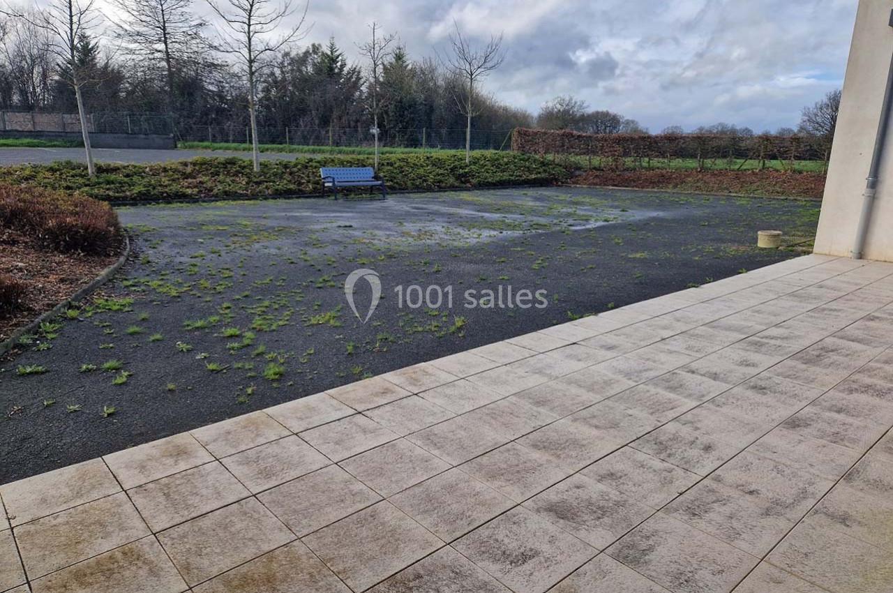 Cour extérieure avec sol en dalles et zone asphaltée partiellement envahie par des herbes, banc bleu en arrière-plan.