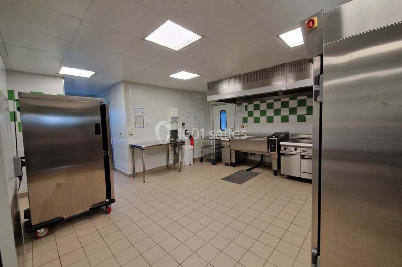 Cuisine professionnelle avec équipements en inox, plan de travail, four, réfrigérateur et carrelage au sol beige.