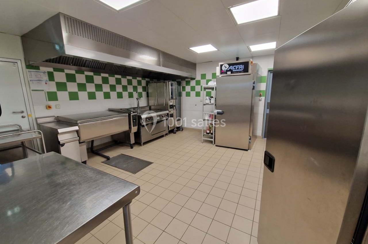 Cuisine professionnelle avec équipements en inox, carrelage beige au sol et murs partiellement carrelés en blanc et vert.