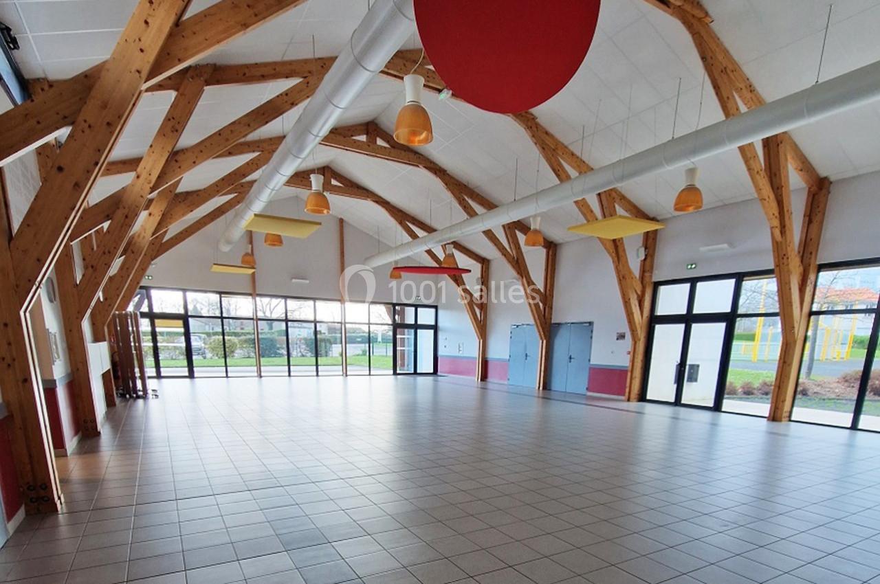 Grande salle lumineuse avec charpente en bois apparente, sol carrelé et larges baies vitrées donnant sur l'extérieur.