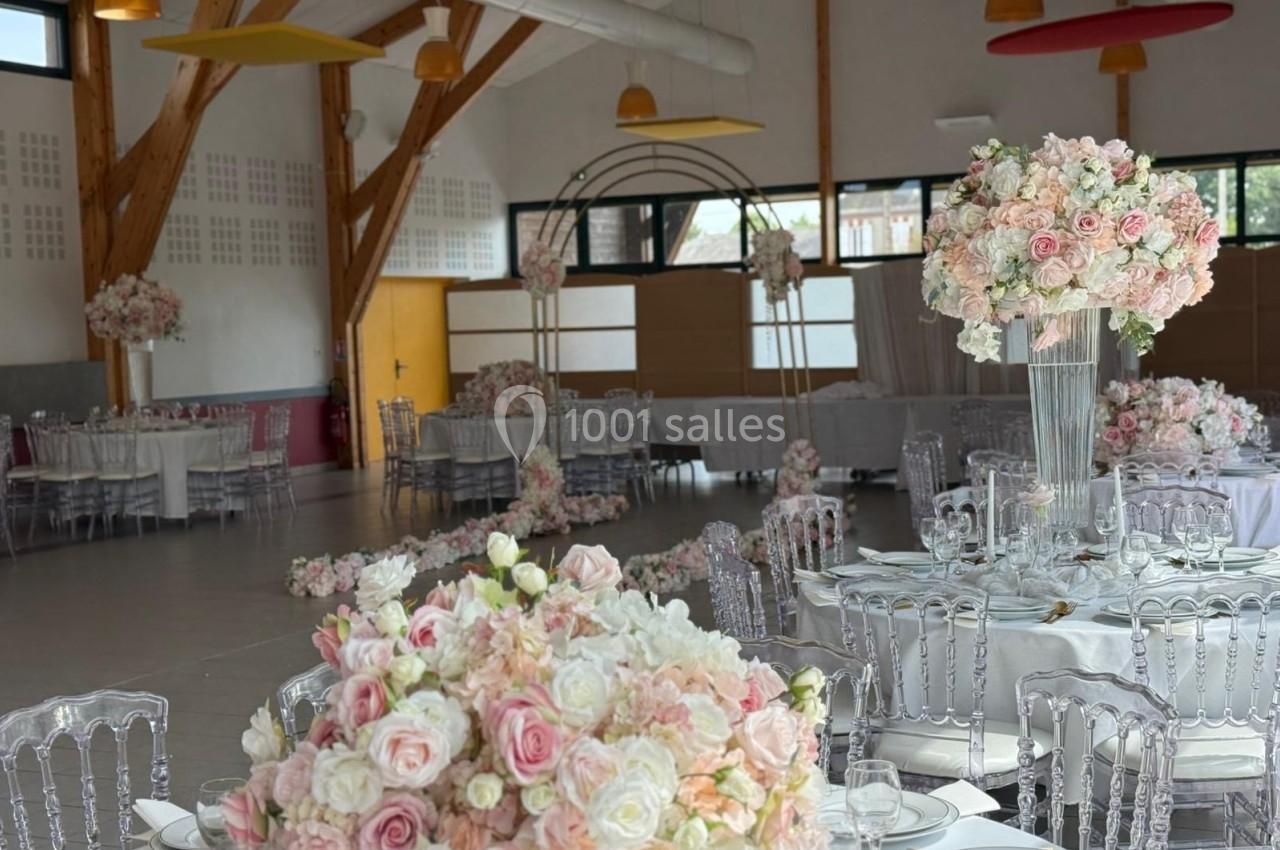 Salle de réception décorée avec des tables rondes, des chaises transparentes et des compositions florales roses et blanches.