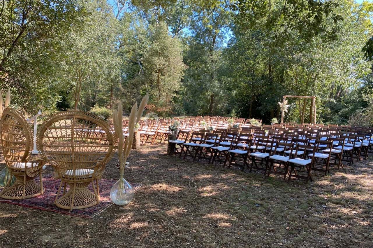 Chaises en bois alignées pour une cérémonie en plein air, avec des fauteuils en rotin et une arche décorative sous les…