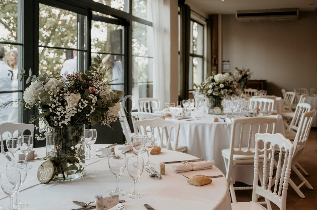 Salle de réception lumineuse avec tables décorées de fleurs, nappes blanches et chaises assorties.