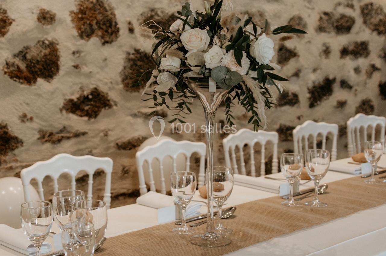 Centre de table floral avec roses blanches sur une table dressée, nappée de beige, devant un mur en pierre.