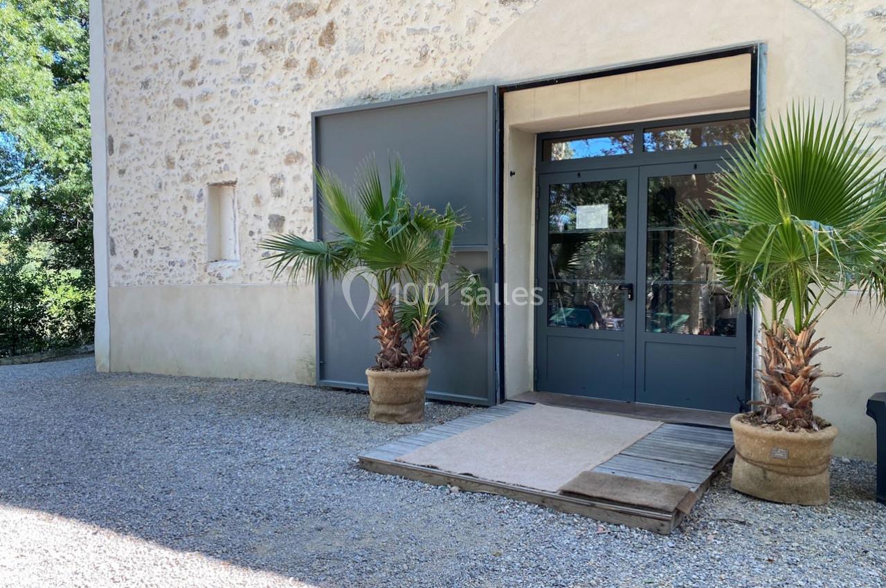Entrée d'un bâtiment en pierre avec une porte vitrée, encadrée par deux palmiers en pot sur un sol gravillonné.