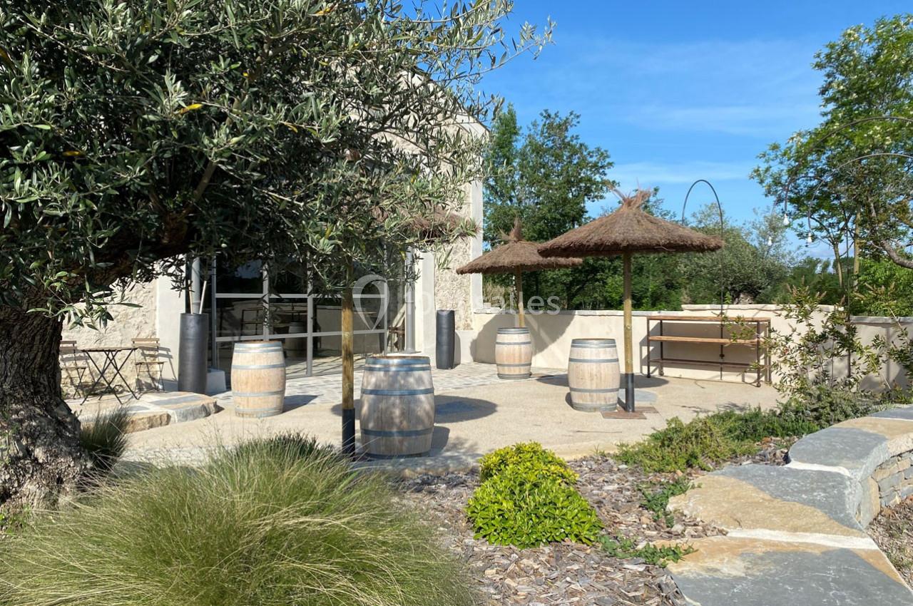Terrasse extérieure avec tables, chaises, tonneaux en bois et parasols en chaume, entourée de végétation méditerranéenne.