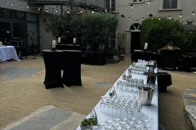 Terrasse aménagée pour un événement avec tables hautes drapées de noir, guirlandes lumineuses et végétation en arrière-plan.