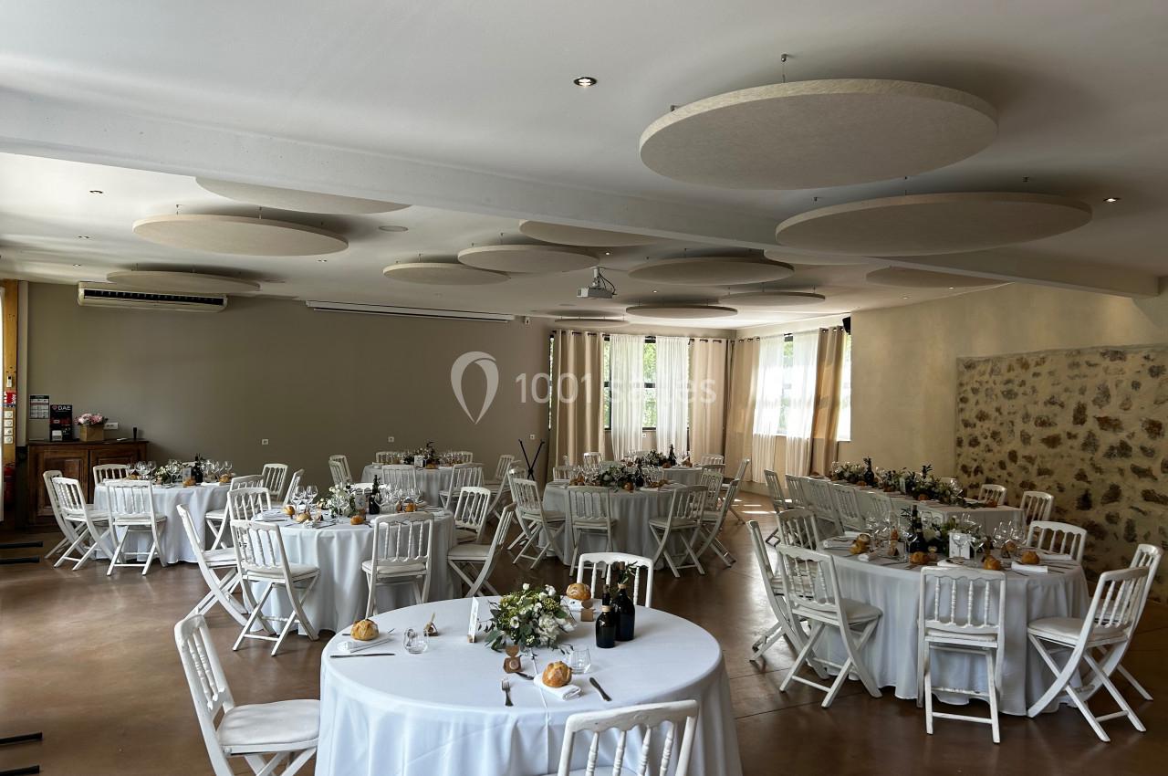Salle de réception décorée avec des tables rondes dressées, nappes blanches, chaises blanches et arrangements floraux.
