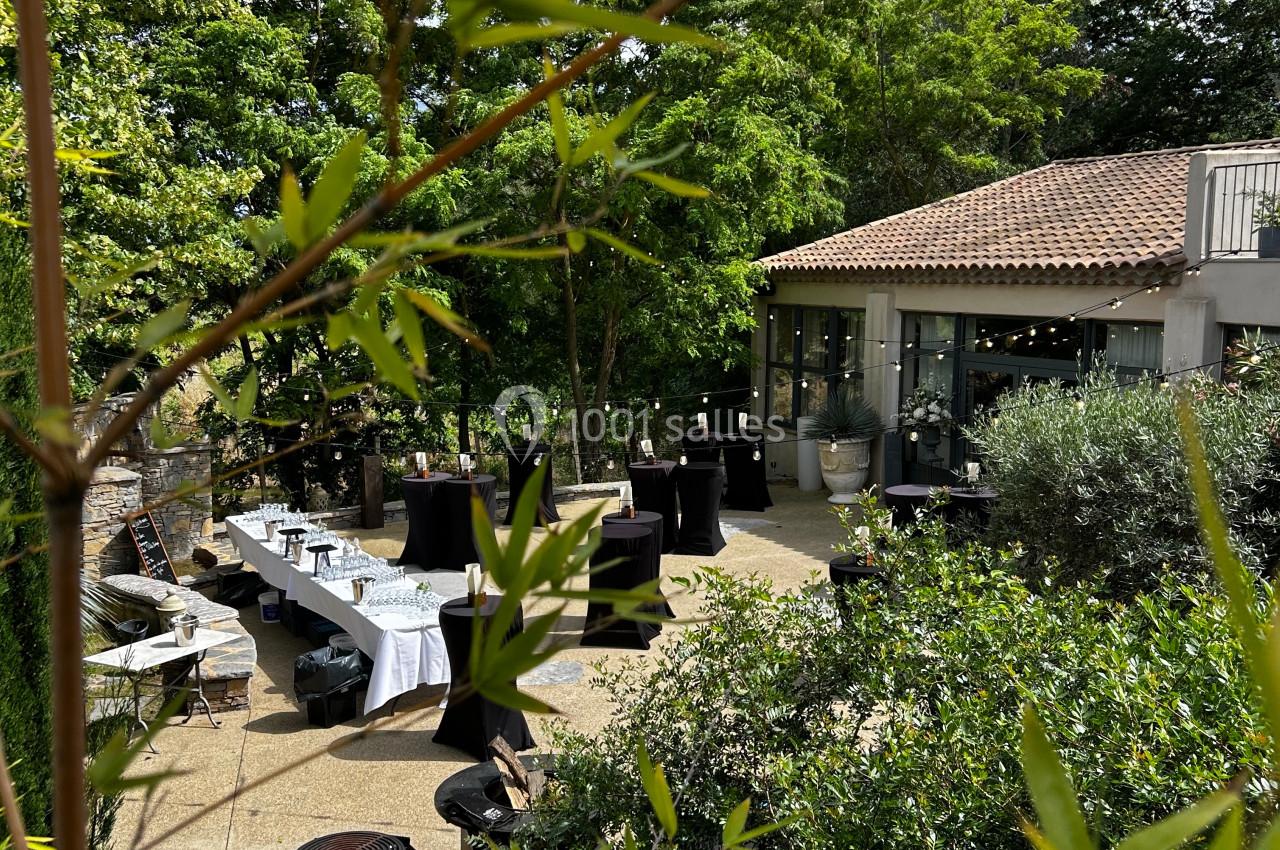 Terrasse aménagée avec tables et chaises, entourée de verdure, devant un bâtiment au toit en tuiles.