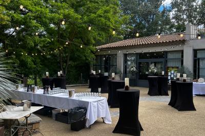 Terrasse aménagée pour un événement avec tables hautes drapées de noir, guirlandes lumineuses et végétation en arrière-plan.