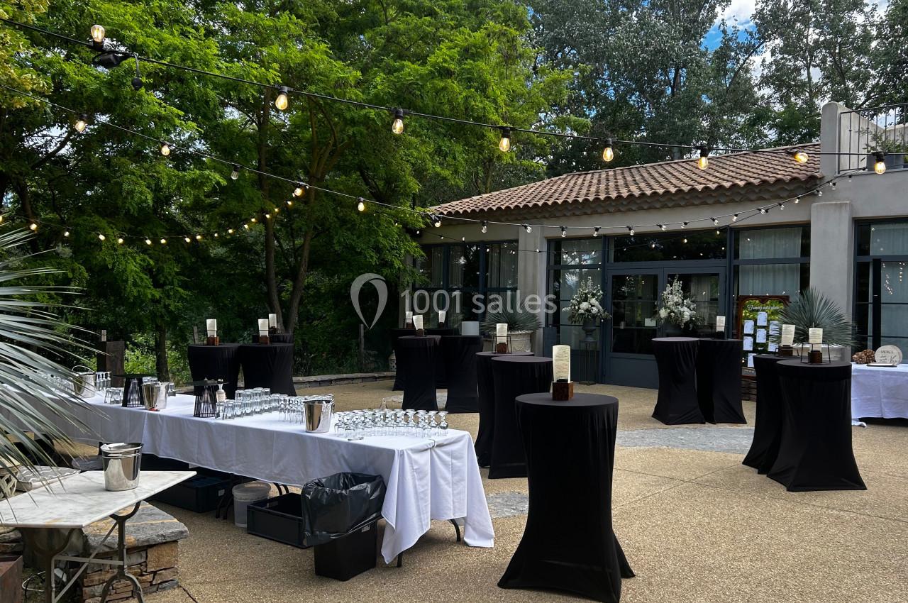 Terrasse aménagée pour un événement avec tables hautes drapées de noir, guirlandes lumineuses et végétation en arrière-plan.