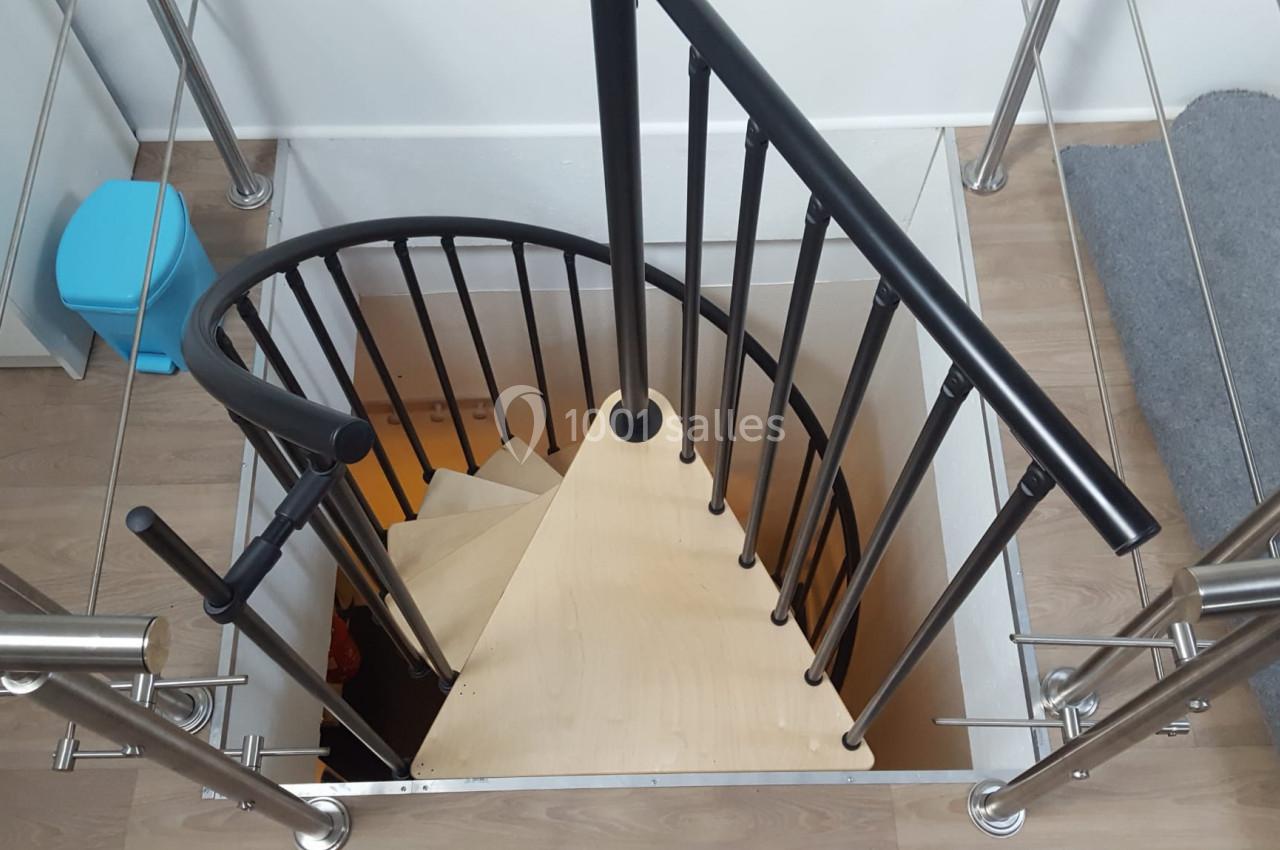 Escalier en colimaçon avec marches en bois clair et garde-corps en métal noir, vu depuis le haut.