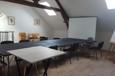 Salle de réunion avec tables noires, chaises, écran de projection, tableau blanc et éclairage naturel.