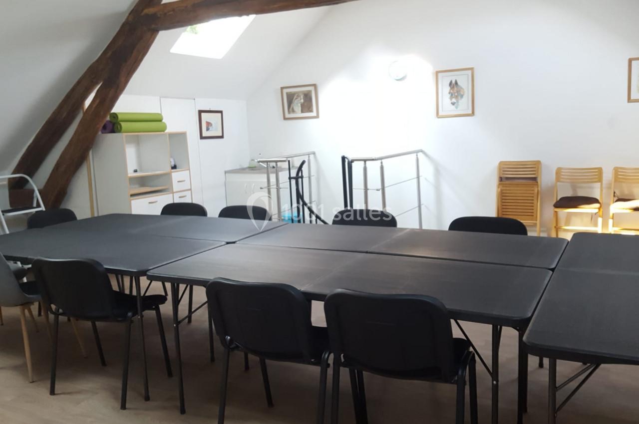 Salle lumineuse avec tables noires disposées en rectangle, chaises autour, étagères et cadres accrochés aux murs blancs.