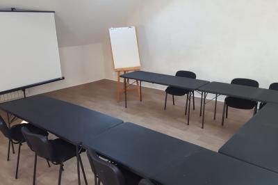 Salle de réunion avec tables noires, chaises, écran de projection, tableau blanc et éclairage naturel.