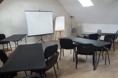 Salle de réunion avec tables noires, chaises, écran de projection, tableau blanc et éclairage naturel.