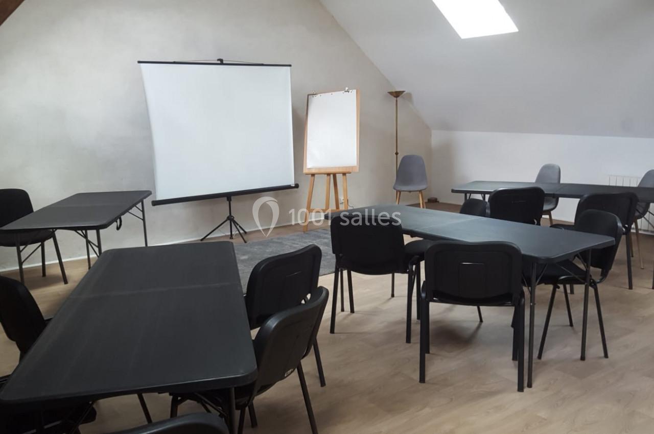 Salle de réunion avec tables noires, chaises, écran de projection, tableau blanc et éclairage naturel.