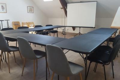 Salle de réunion avec tables noires, chaises, écran de projection, tableau blanc et éclairage naturel.