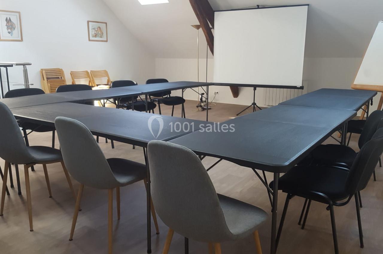 Salle de réunion lumineuse avec tables disposées en U, chaises, tableau blanc et écran de projection.