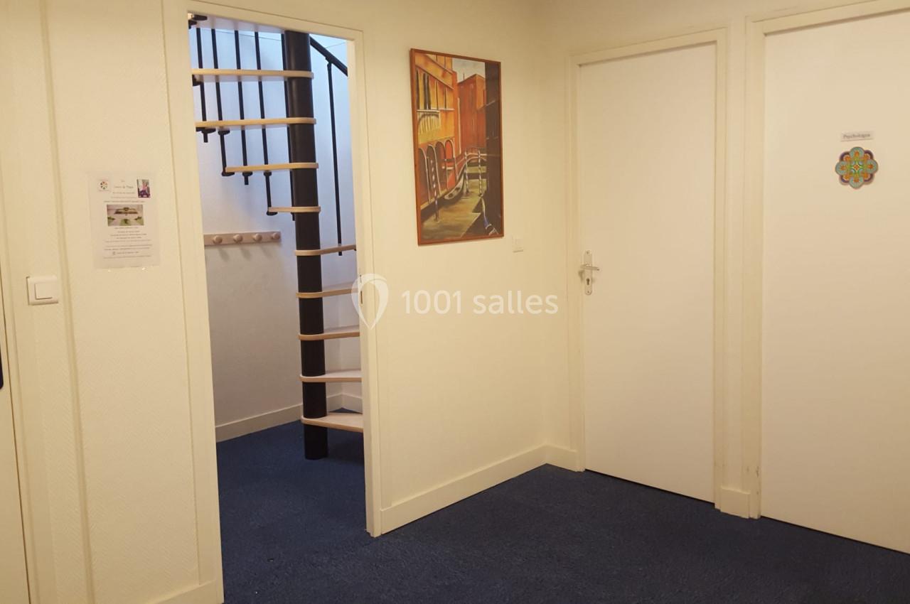 Couloir avec moquette bleue, deux portes blanches fermées, un escalier en colimaçon et un tableau mural coloré.