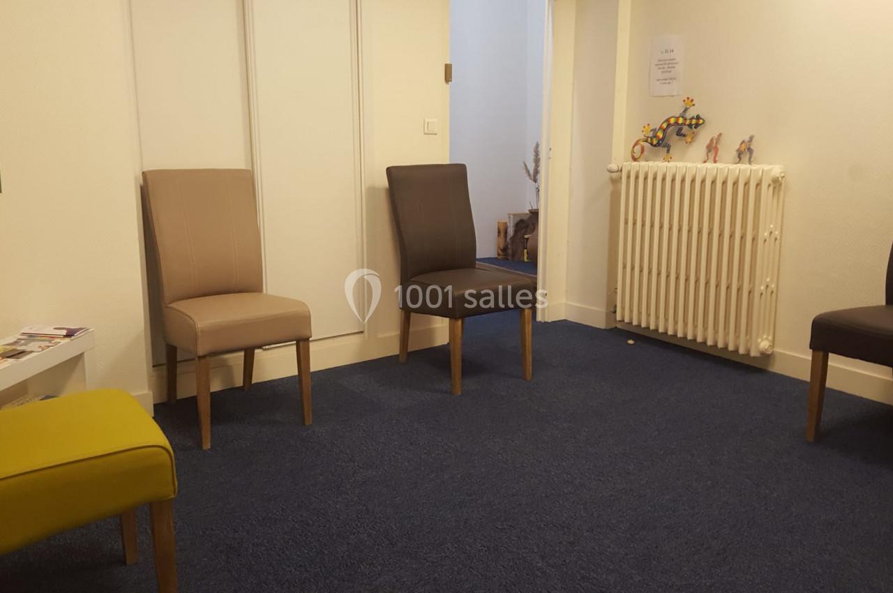 Salle d'attente avec quatre chaises disposées autour d'un radiateur, sol en moquette bleue et murs clairs.