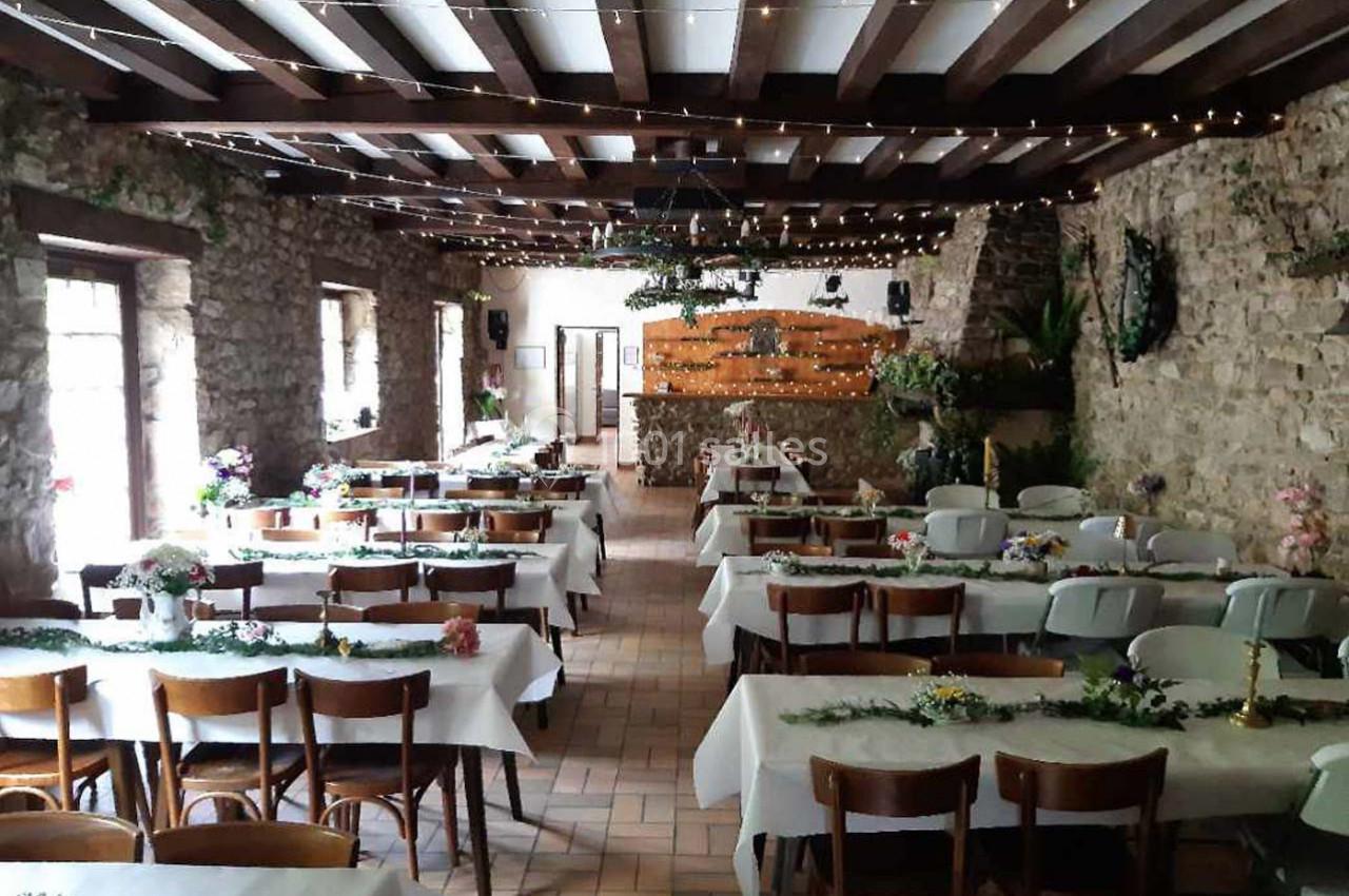 Salle de réception rustique avec murs en pierre, tables décorées de nappes blanches et guirlandes lumineuses au plafond.