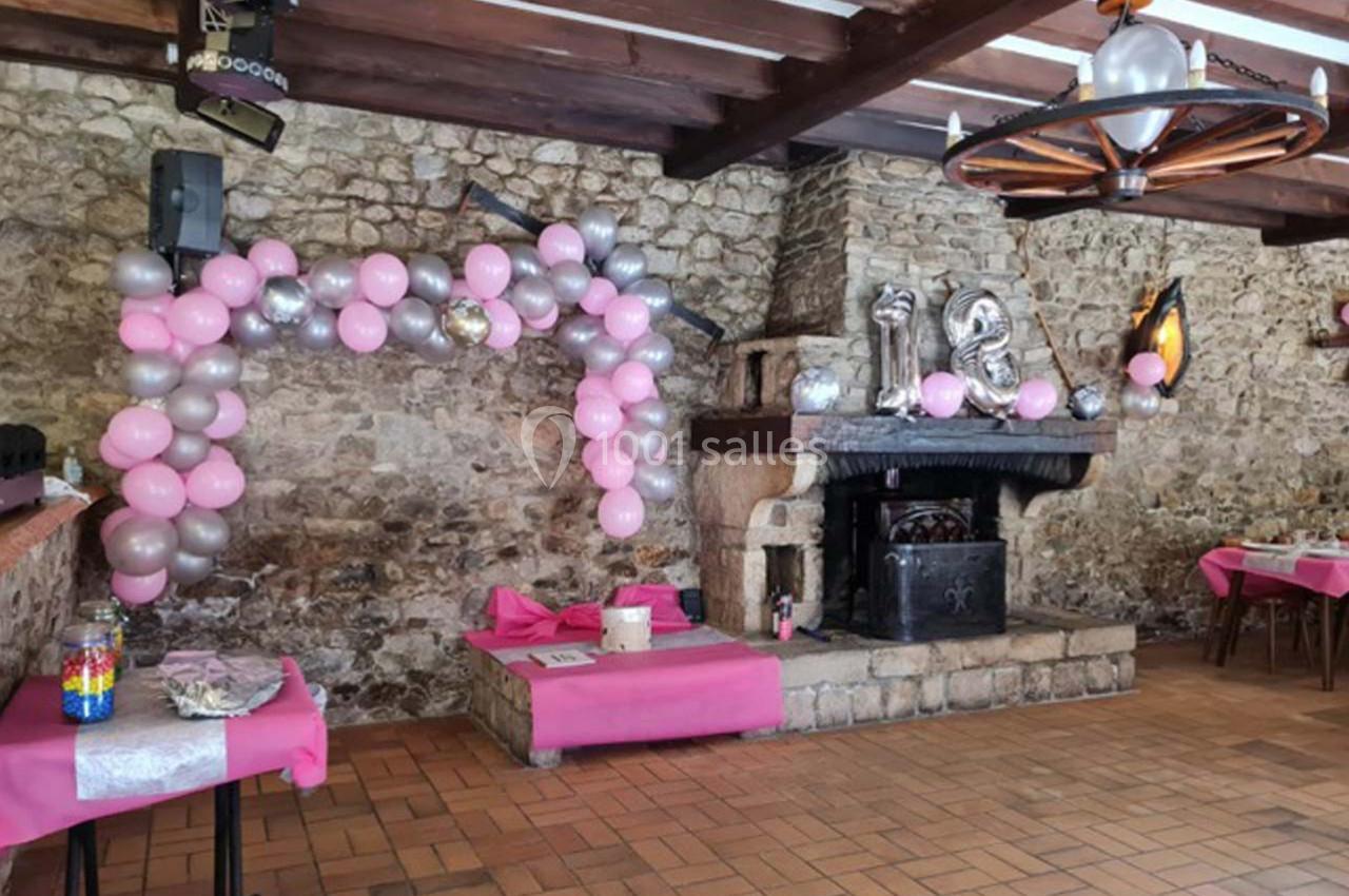 Salle décorée pour une fête, avec des ballons roses et argentés près d'une cheminée en pierre.