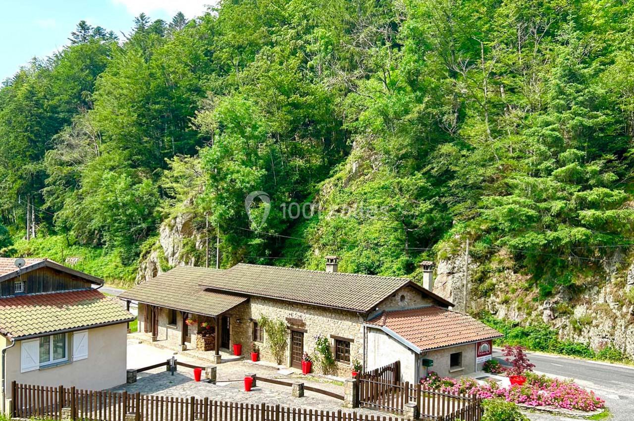 Bâtiments en pierre et en bois entourés de végétation dense, situés près d'une route et d'une colline boisée.