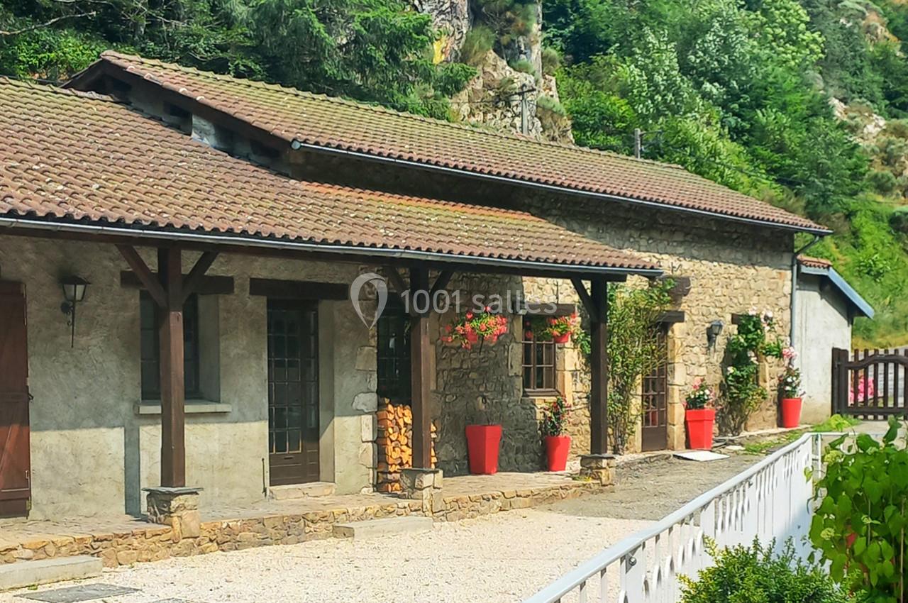 Façade d'une maison en pierre avec toit en tuiles, pots de fleurs rouges et végétation environnante.