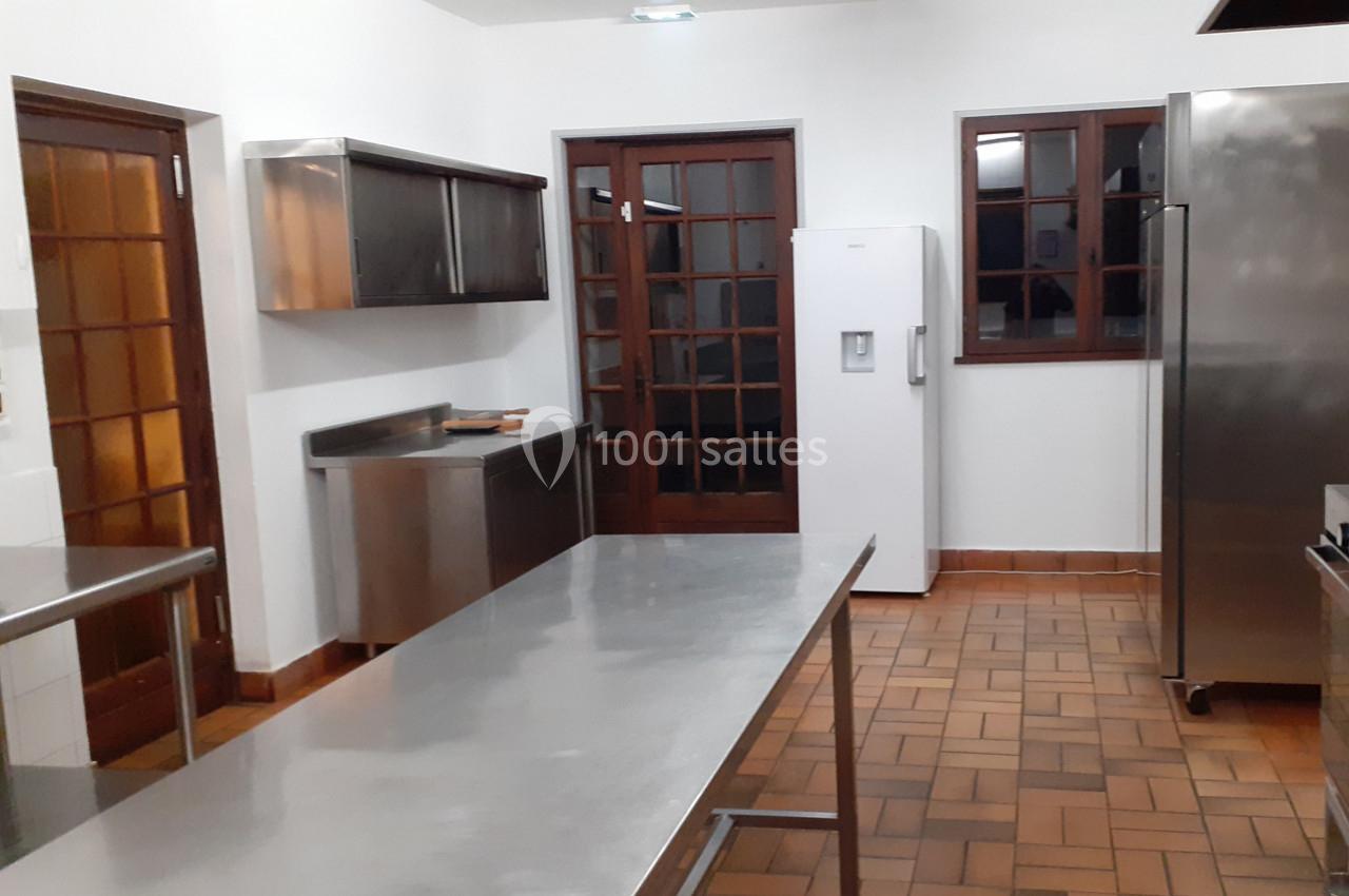 Cuisine professionnelle avec plans de travail en inox, réfrigérateur blanc, étagères murales et sol en carrelage.