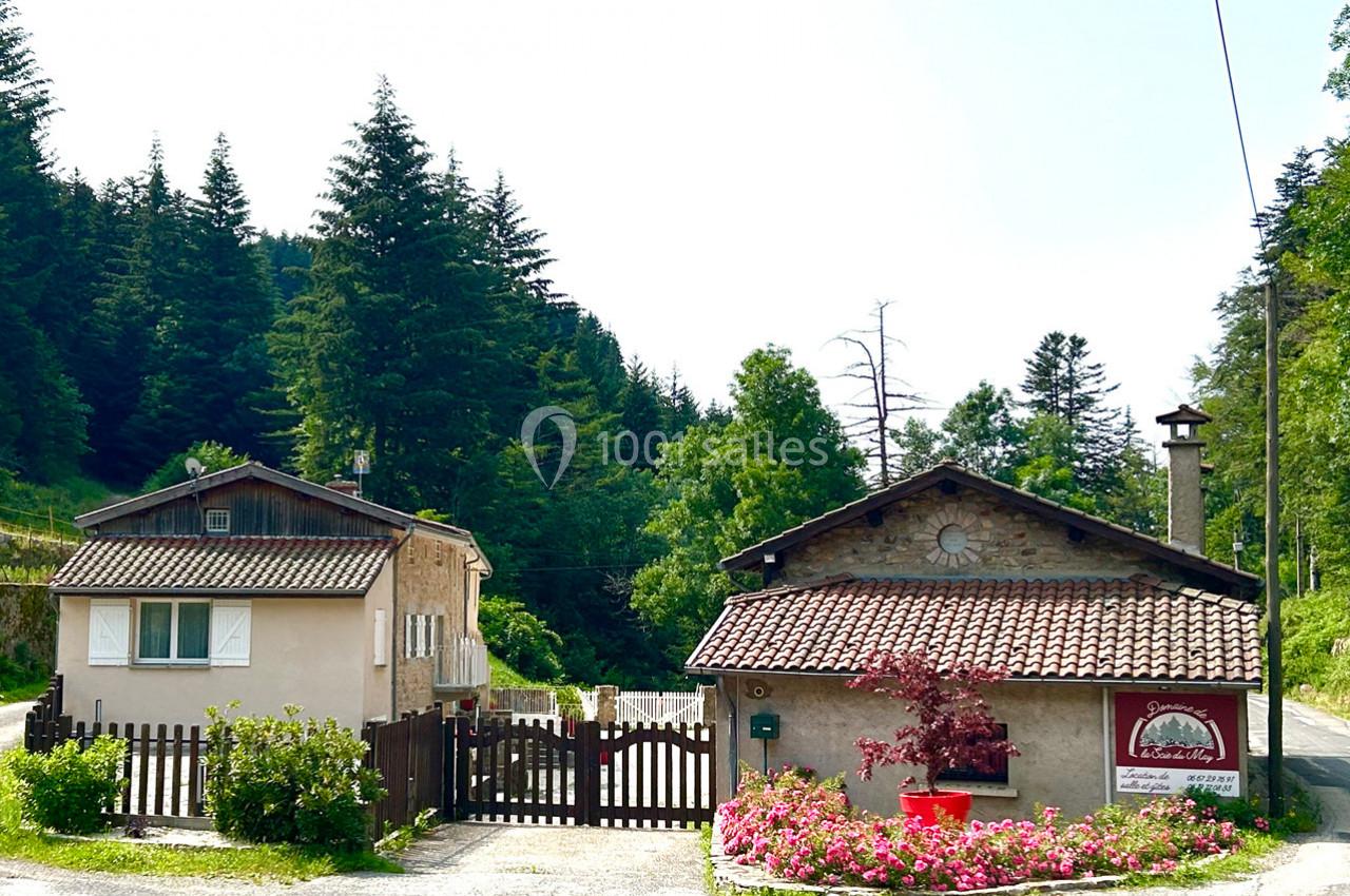 Deux maisons rurales avec toits en tuiles, entourées de végétation et de fleurs, bordant une route forestière.
