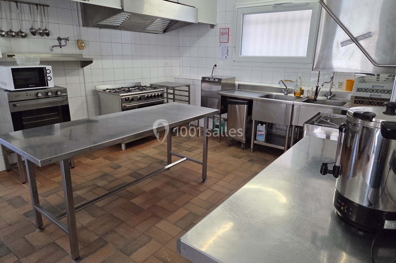 Cuisine professionnelle équipée avec plans de travail en inox, appareils de cuisson et évier sur carrelage brun.