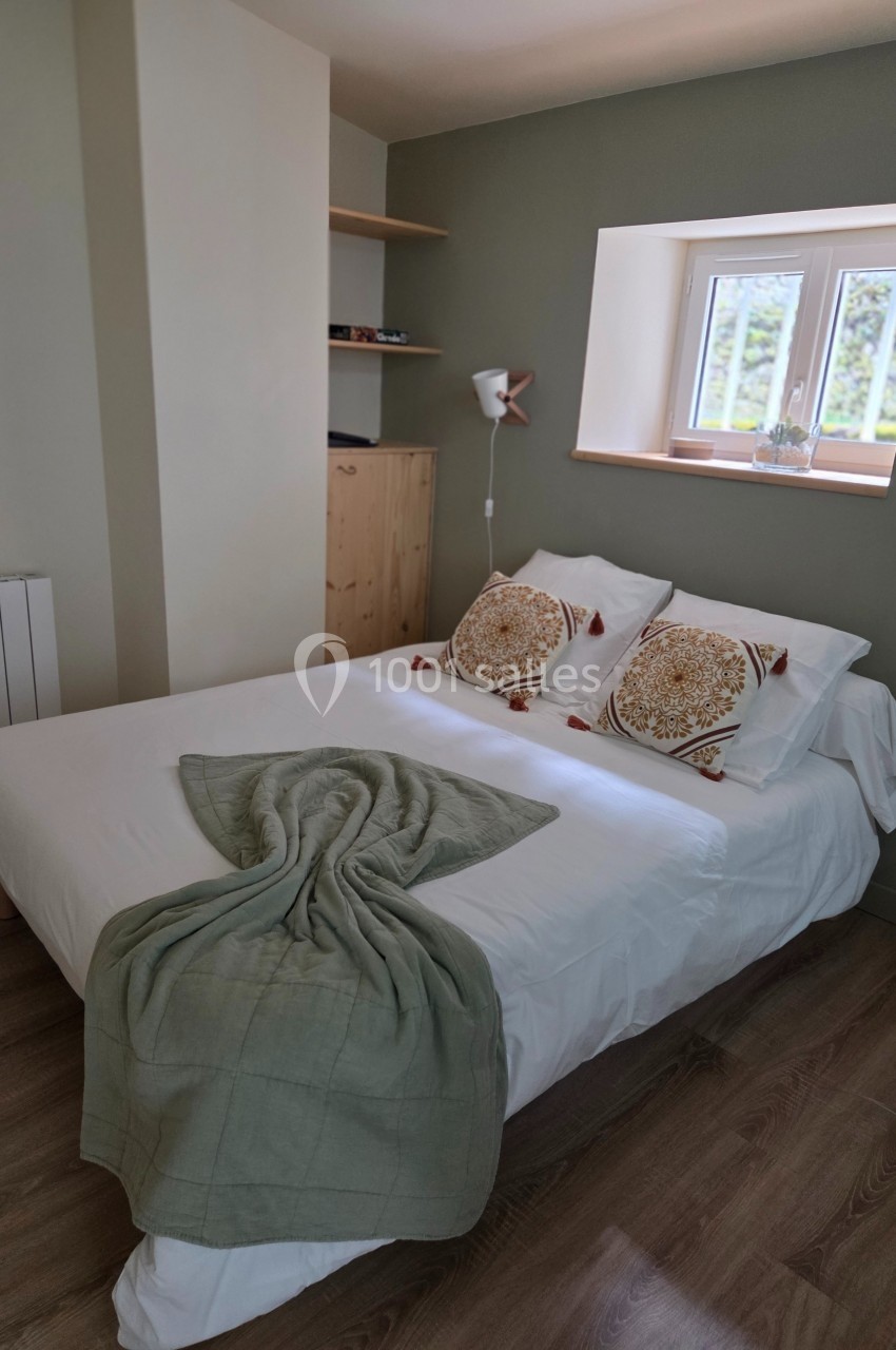 Chambre lumineuse avec lit double, plaid vert plié, coussins décoratifs et fenêtre donnant sur un extérieur verdoyant.