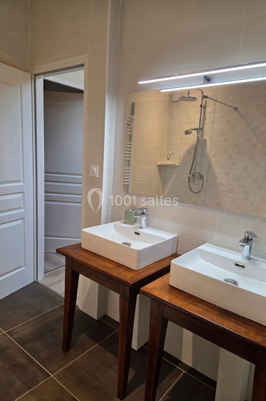 Salle de bain moderne avec deux vasques sur des meubles en bois, miroir éclairé et douche à l'arrière-plan.