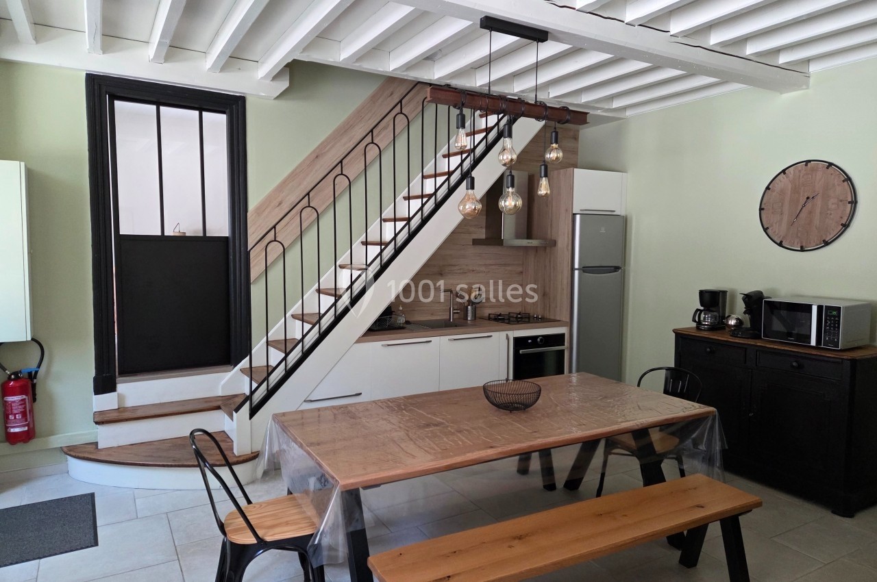 Cuisine moderne avec table en bois, chaises métalliques, escalier en bois, et électroménagers sous un plafond à poutres…