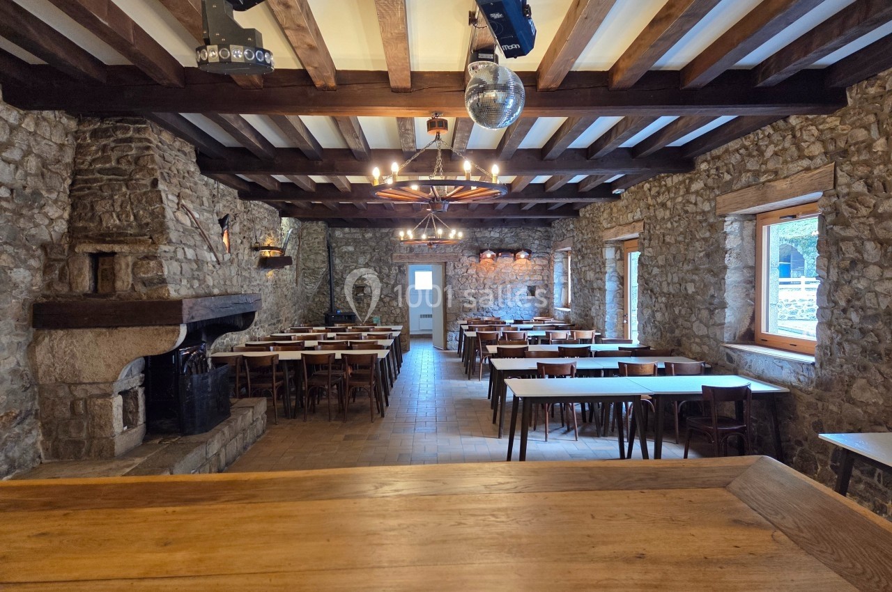 Salle rustique avec murs en pierre, poutres apparentes, cheminée, tables alignées et grandes fenêtres.