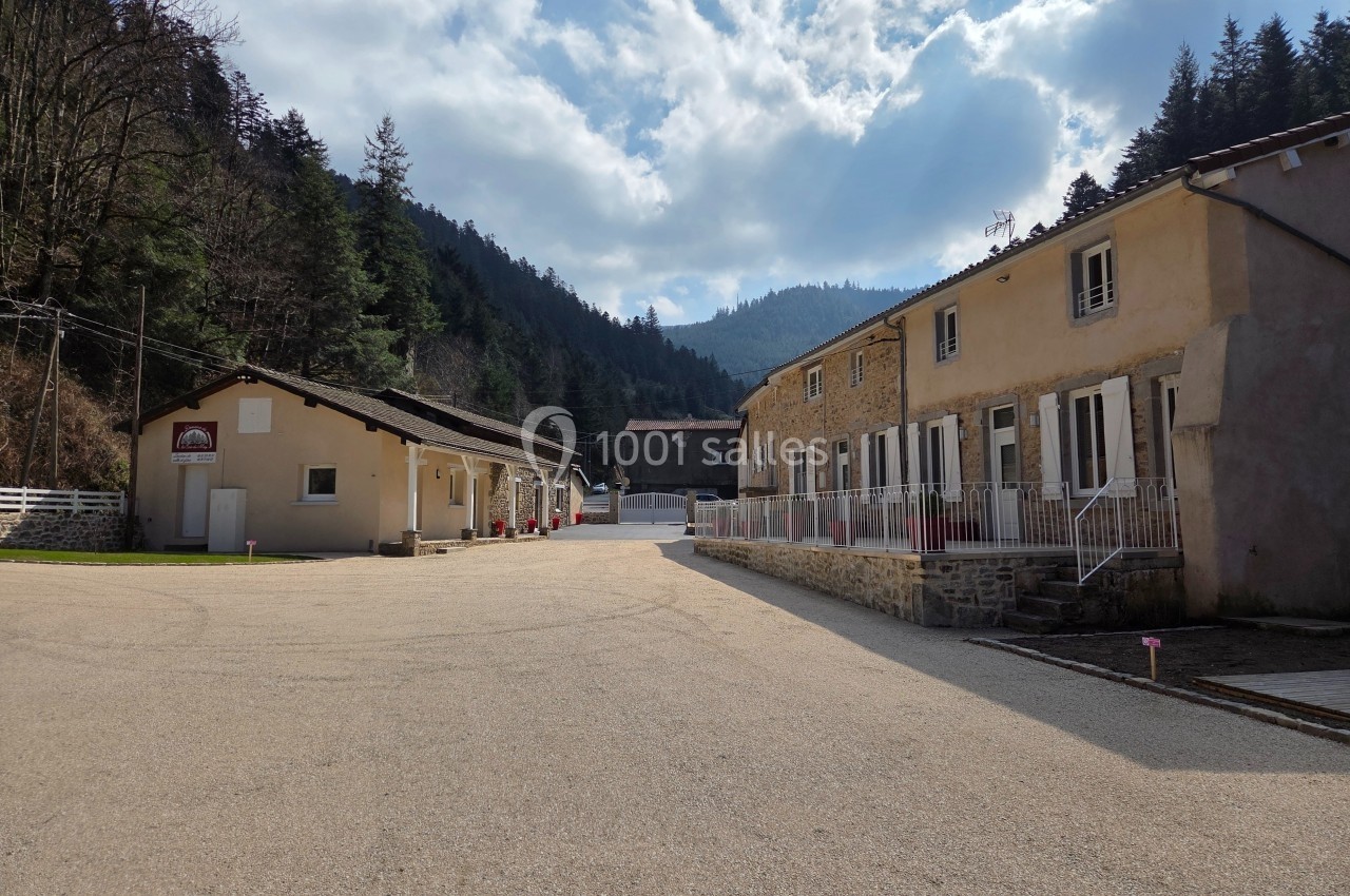 Cour d'un ensemble de bâtiments en pierre et crépi, entourée de collines boisées sous un ciel partiellement nuageux.