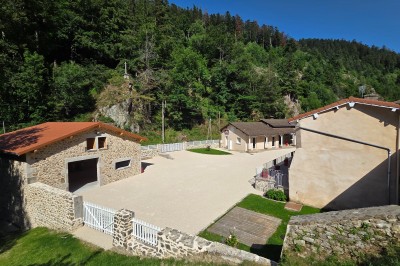 Façade d'une maison en pierre avec toit en tuiles, entourée de verdure et d'une allée asphaltée.