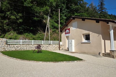Façade d'une maison en pierre avec toit en tuiles, entourée de verdure et d'une allée asphaltée.