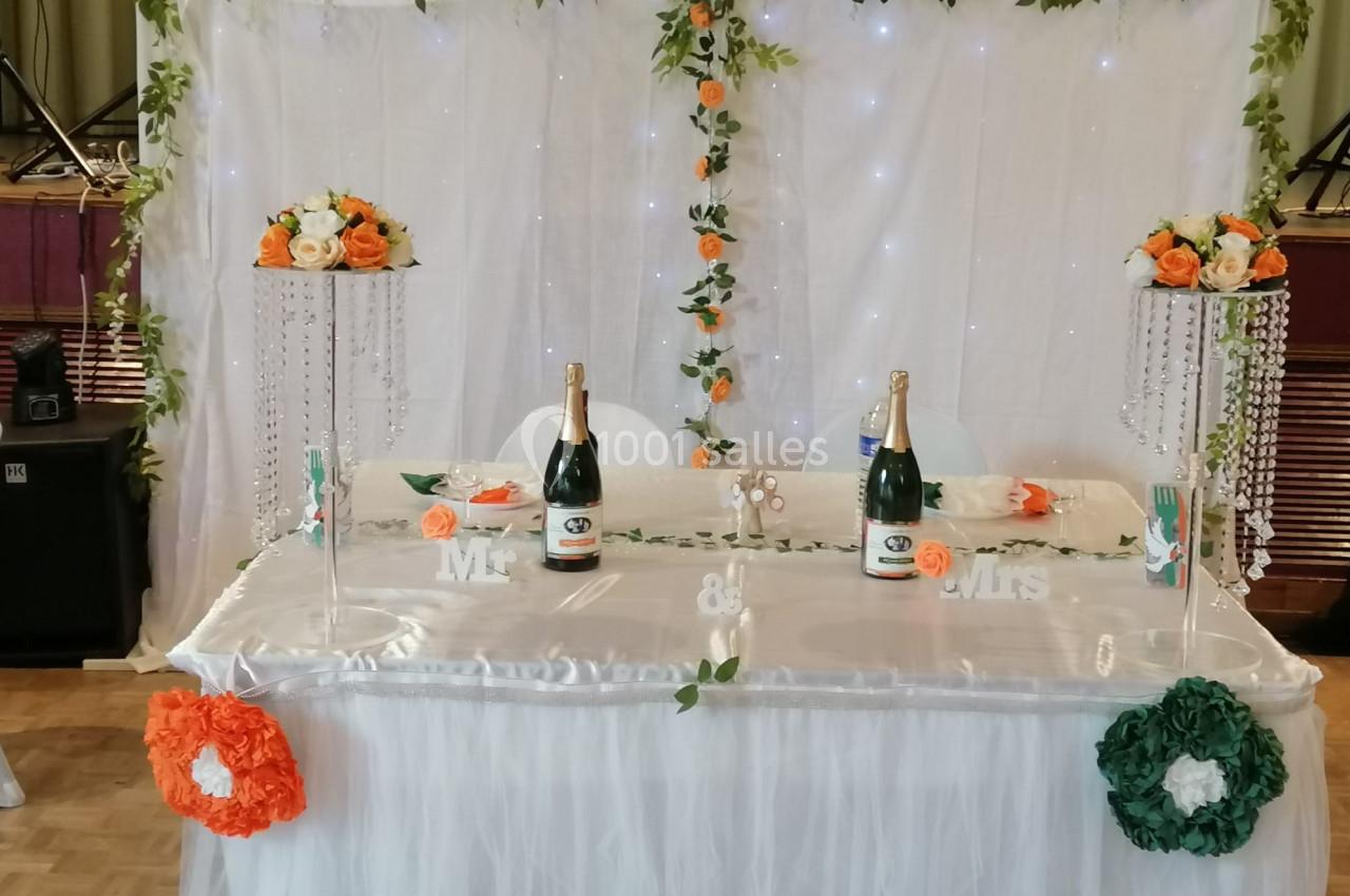Table décorée pour un mariage avec nappes blanches, fleurs orange et blanches, bouteilles de champagne et guirlandes…