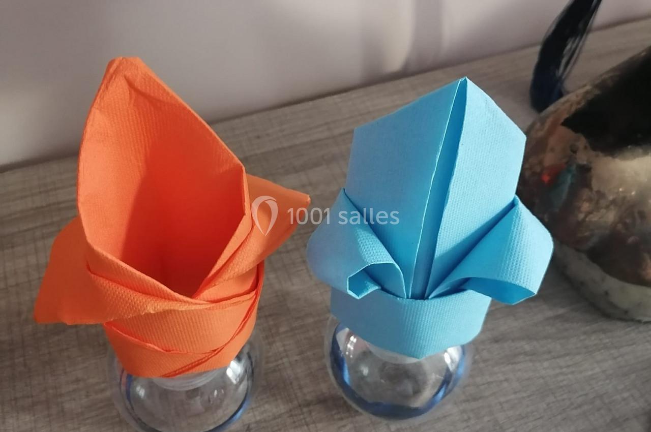 Serviettes pliées en forme de fleurs, orange et bleue, disposées sur des verres transparents sur une table en bois.
