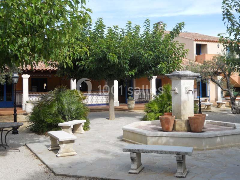 Cour intérieure avec fontaine centrale, bancs en pierre, arbres et bâtiment aux murs ocres et volets bleus en arrière-plan.